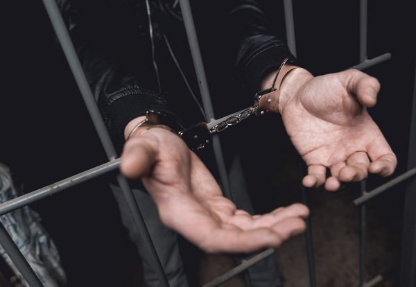 Man In Handcuffs Behind Bars In A Police Station.